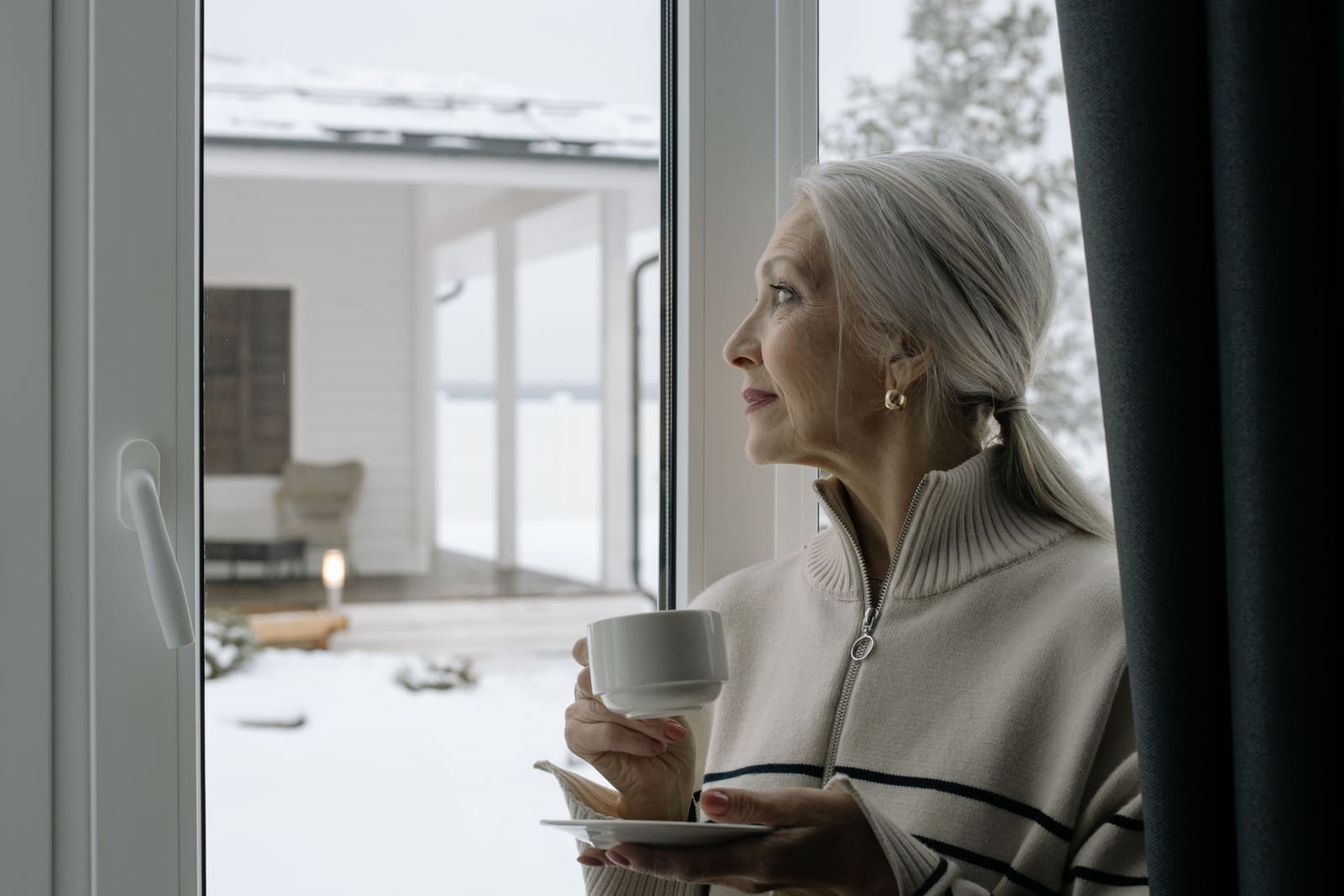 Woman in a knit sweater holding a cup of coffee and looking out a window at a snowy yard